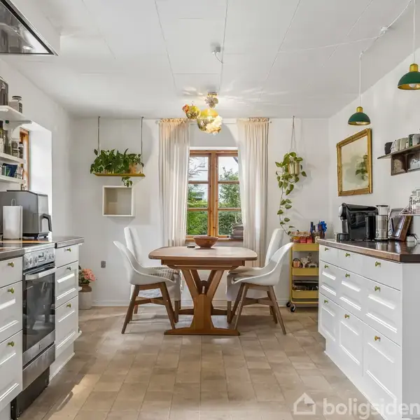 Et køkken med hvide skabe og træbord. Bordet har fire stole omkring det. Grønne planter og bøger dekorerer rummet, og to vinduer lader naturligt lys komme ind.