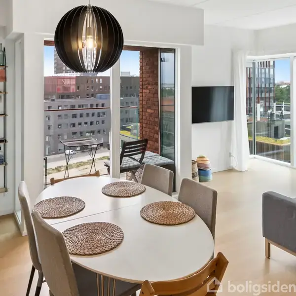 Et lyst rum med et ovalt spisebord, dækket med runde bordskånere, står i midten. Ved siden af er en grå sofa og en stor lampe hænger over bordet. Balkon med udsigt over bybygninger i baggrunden. En lille reol, væghængt tv, og døre til altan ses også.