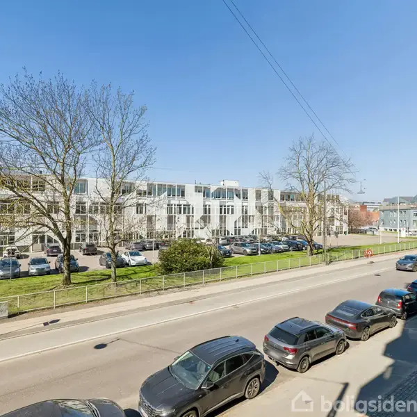Et gadebillede med parkerede biler langs fortovet. Baggrunden viser et stort, hvidt byggeri med mange vinduer, omgivet af grønne områder og træer under en klar blå himmel.