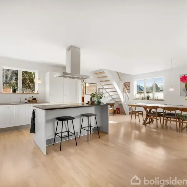 Køkken med kogeø og barstole. Åben planløsning med spisebordsområde, lyse trægulve, trappe til øverste etage og store vinduer med udsigt. Dekoreret med planter og malerier.
