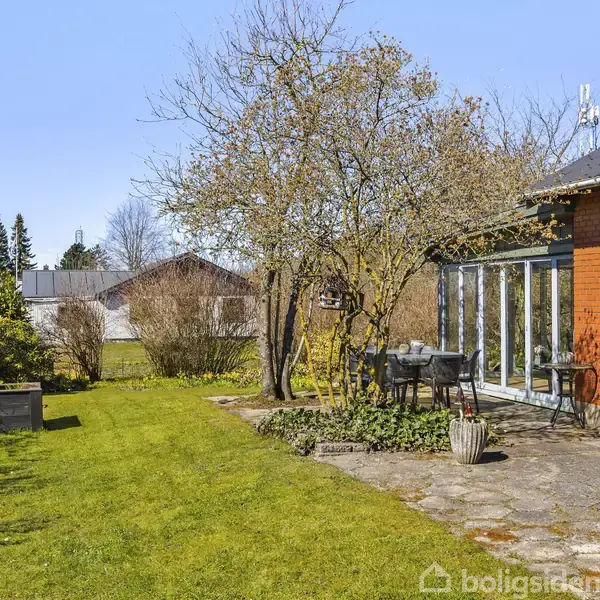 Rødt murstenshus med store vinduer, omkranset af frodig have med græsplæne og blomstrende træer. Et udendørs siddeområde med bord og stole ligger ved siden af huset.