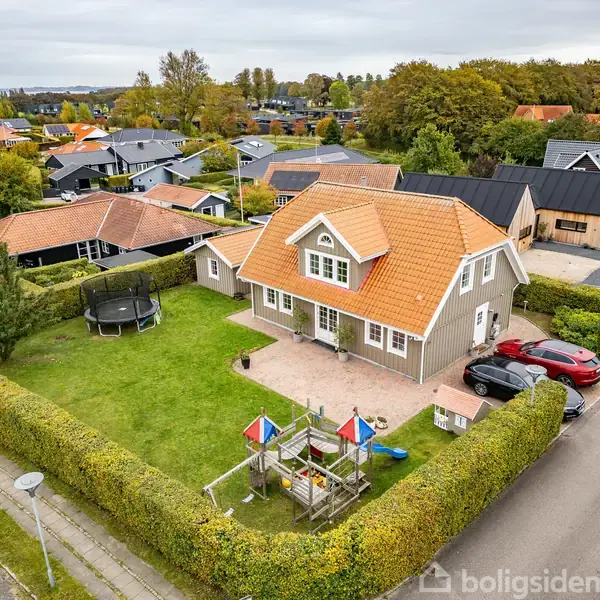 Huset står på en velholdt grund med grøn plæne og buskhegn. I haven er der en trampolin og en legeplads. Omgivelserne er et villakvarter med flere huse i baggrunden.