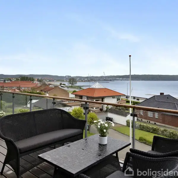 Terrassemøbler står på en altan med glasværn, der tilbyder udsigt over en by med huse og en fjord. På bordet står blomster i potter som dekoration.
