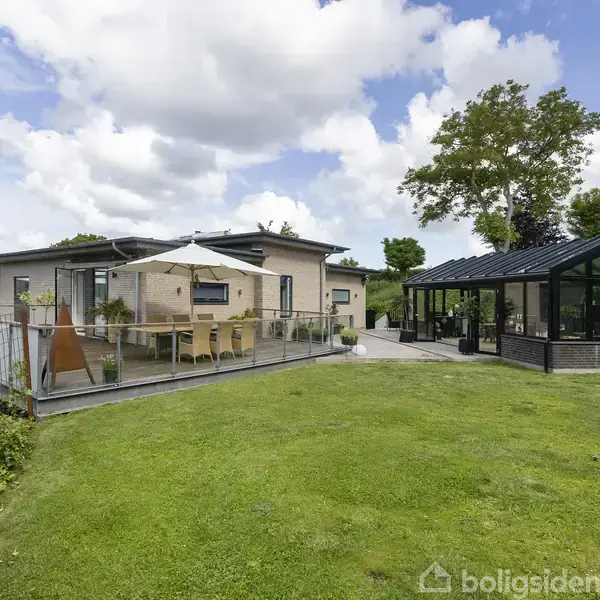 Hus med terrassebord og parasol står ved siden af en udestue med glasvægge. Græsplæne og træer danner baggrund med udsigt over vandet under en skyet himmel.