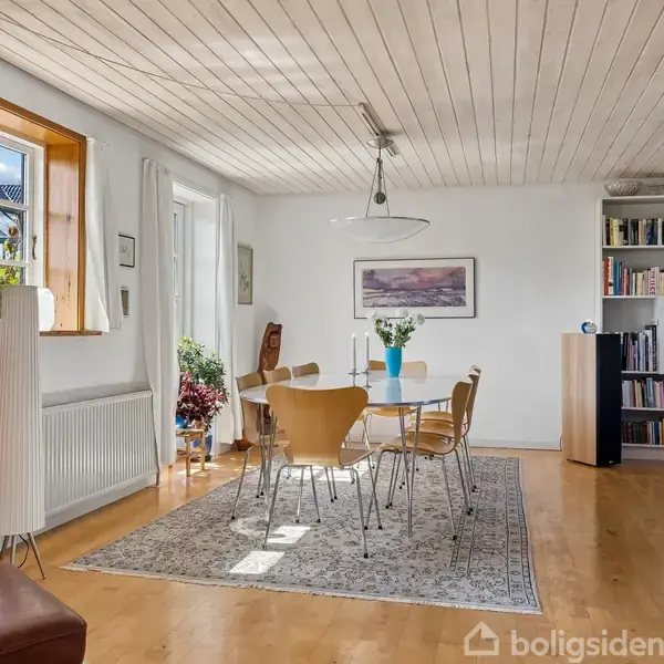 I et lyst spisestue sidder seks stole omkring et bord med en vase. Rummet har træloft, et stort vindue, reol og dekorationer, der skaber en hyggelig atmosfære.