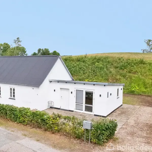 Hvidt hus med sort tag står stille ved en sti, omgivet af frodige grønne marker og buske, under en klar blå himmel.