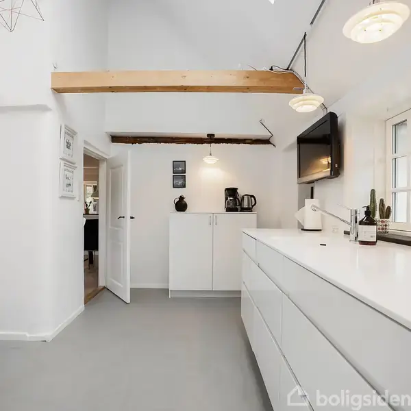 Køkken med hvide skabe og skuffer under en trækonstruktion. Der står en kaffemaskine og en kande på et skab. Der hænger en fladskærm over bordpladen ved vinduet.