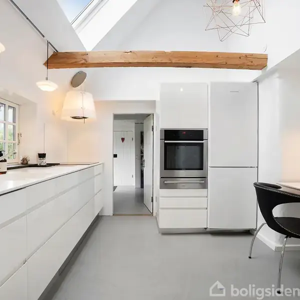 Køkkenet har hvide skabe og en indbygget ovn; en sort stol står ved et lille bord. Ovenlys og træbjælke i loftet skaber en lys atmosfære.
