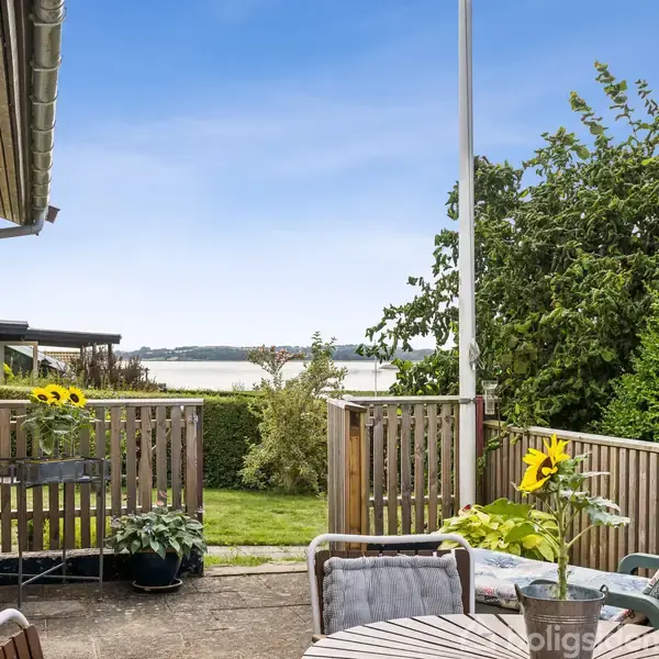 Terrasse med runde stole omkring et bord med solsikkeblomster. Handlingen er hvilestunder på en solrig dag. Omgivelserne inkluderer have og søudsigt med blå himmel.