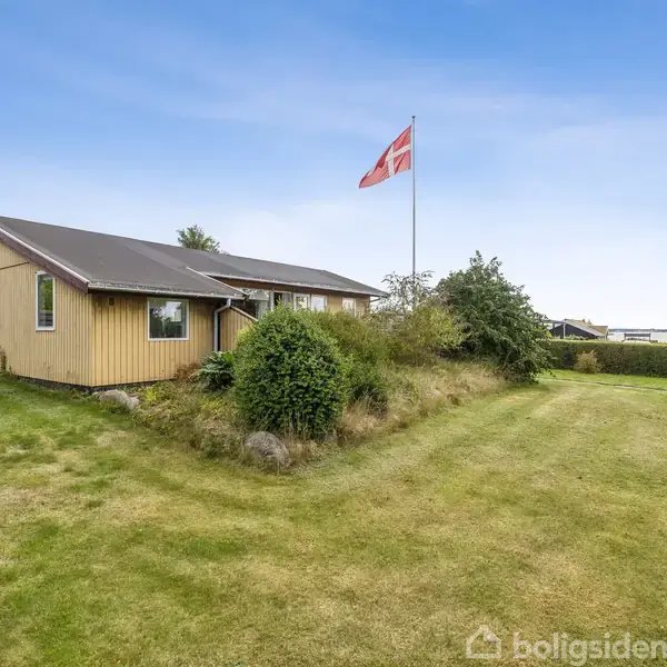 Et gult hus står stille i en grøn have under en klar blå himmel, med et dansk flag vajende fra en flagstang, omgivet af frodige træer og buske.