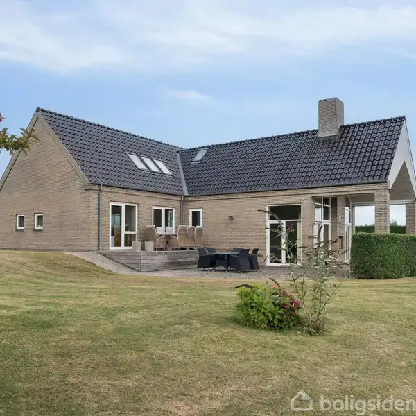 Et moderne hus med gråt tag og store vinduer står i en velplejet have. Der er en terrasse med havemøbler, omgivet af græsplæne.