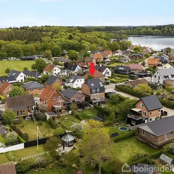 Rødt hus markeret med pil står blandt flere huse i en frodig, grønt beboelsesområde med træer og en sø i baggrunden.