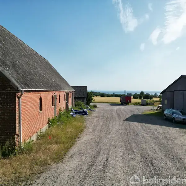 En rød murstenslade står stille på en gårdsplads med grus. En bil og en trailer findes i nærheden, omgivet af åbne marker og en klar blå himmel.
