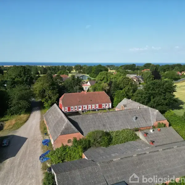 Rødt murstenshus står omgivet af gårdsbygninger og en grusvej, landskab af skov og kornmarker med havet i baggrunden under en klar blå himmel.