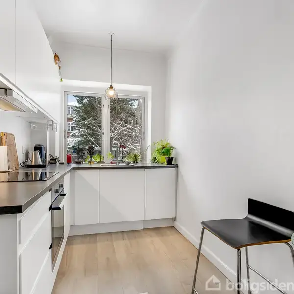 Et moderne køkken med hvide skabe og sort bordplade. Køkkenet har en komfur og vask. Et vindue med udsigt til træer og flere planter på vindueskarmen. En sort stol står til højre.