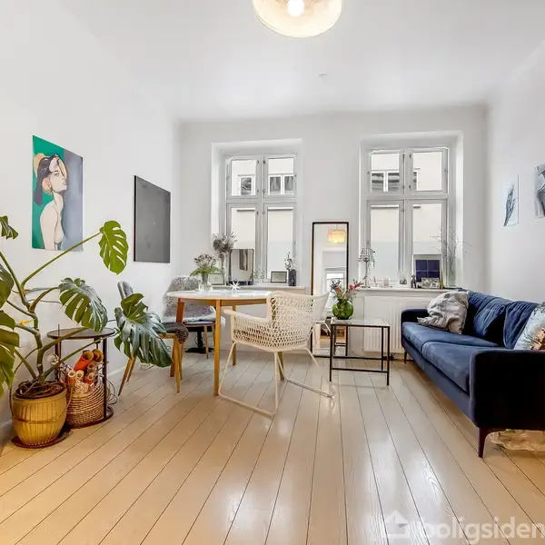 En lys stue med en blå sofa til højre, to store vinduer bagved, og planter i venstre hjørne. Et spisebord med stole står foran vinduerne, omgivet af kunstværker.