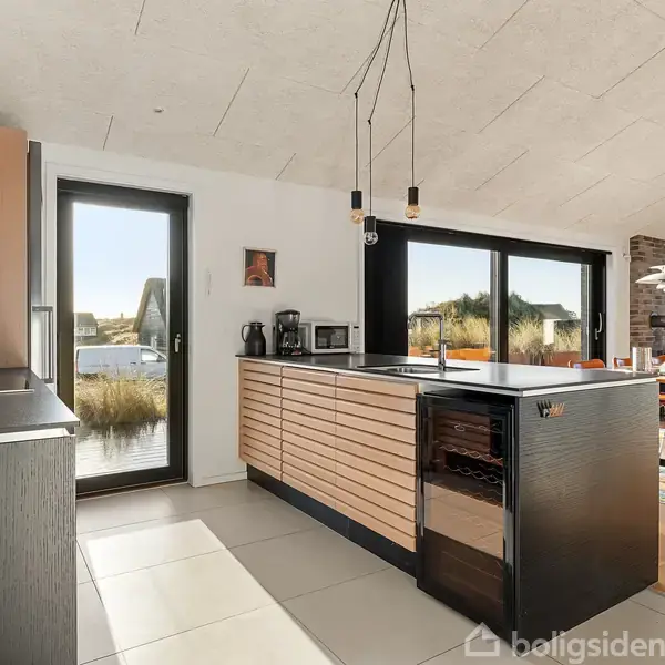 Køkken med træpaneler på en ø, kaffemaskine og brødrister står på bordet. Stort vindue og dør med udsigt til have, spisebord i baggrunden.