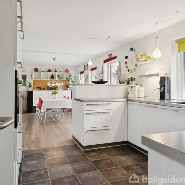 Køkken i moderne stil har hvide skabe og bordplader. Det er veloplyst med vindue mod højre og hængende lamper. Åben forbindelse til spisestue set i baggrunden.