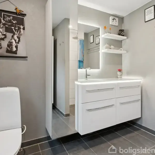 Hvidt badeværelsesmøbel med vask og skuffer, placeret i et gråt badeværelse. Til venstre en toiletstol. På væggen hænger indrammede billeder og en hylde med rengøringsartikler.