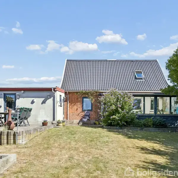 Et moderne hus med både murstens- og træfacade står i en solrig have. Terrassen til venstre har havemøbler og en parasol. Blå himmel med få skyer.