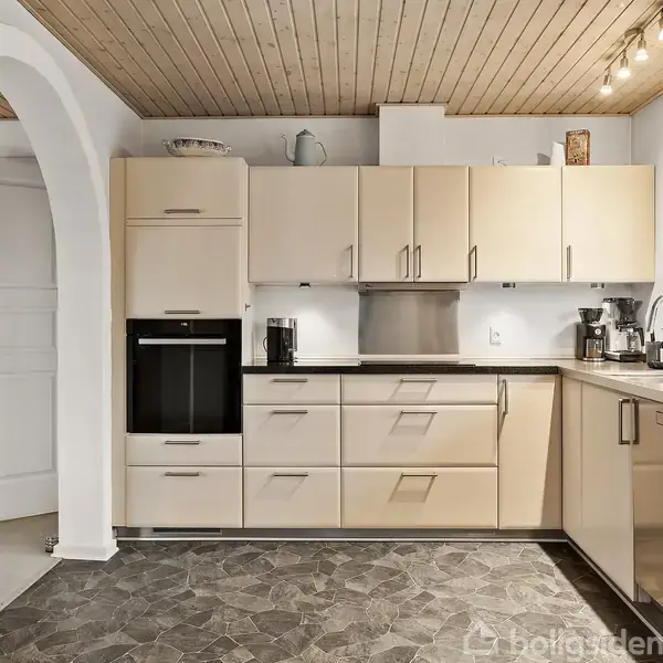 Køkken med lyse skabe og sorte bordplader, rummer en indbygget ovn og kaffemaskine. Et firkantet vindue lader lys ind, mens gulvet har et mørkt mønster.