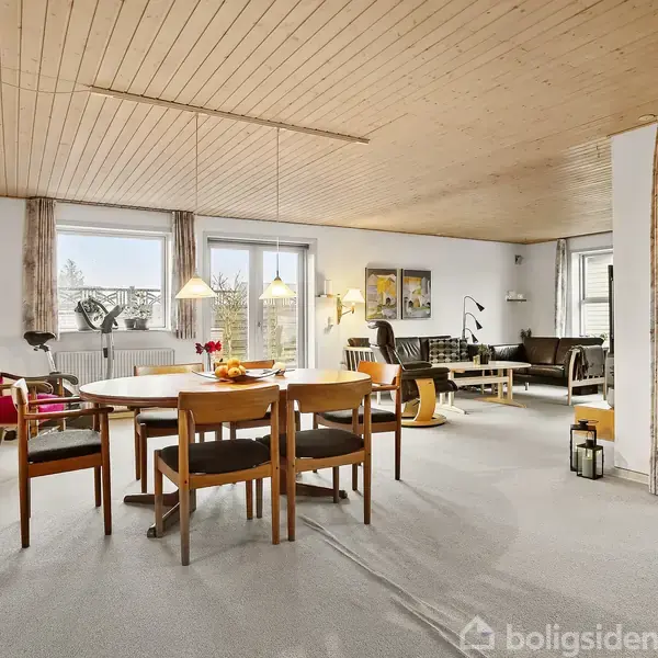 Spisebord med stole står i en stue. Det er oplyst af hængelamper. Stuen har lyse vægge, træloft og sofaområde med store vinduer, som giver udsigt til en have.