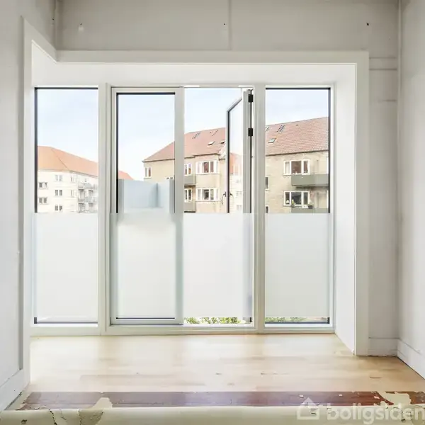 Glassdør står åben mod en balkon; udsigt over boligbygninger med røde tage ses igennem døren. Indendørs, rummet er lyst med lyse trægulve og hvide vægge.