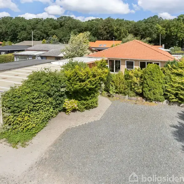 En villa med rødt tag står omgivet af buske og træer i et boligområde. En grusbelagt indkørsel leder til en carport. I baggrunden ses flere træer.