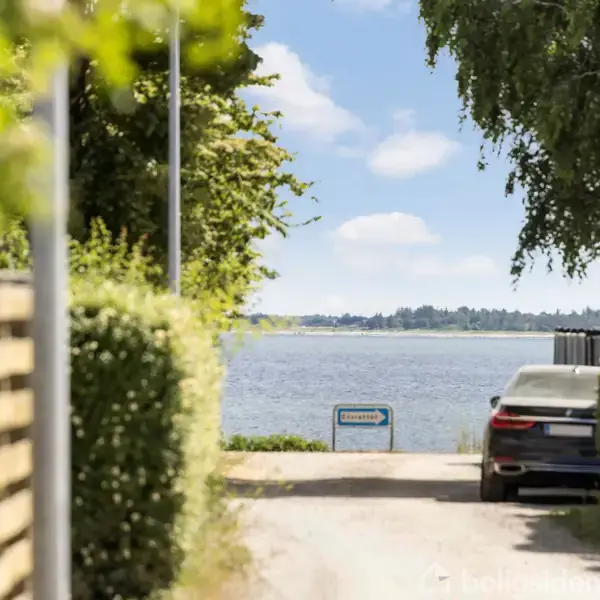 En bil holder parkeret på en smal vej omkranset af grønne hække. I baggrunden ses en udsigt over vandet og et skilt med teksten "Fjordstien".