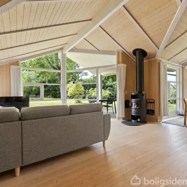 Gråt sofaarrangement står i en stue med træbeklædte vægge og loft, hvor en moderne brændeovn er centrum. Store vinduer giver udsigt til en grøn have.