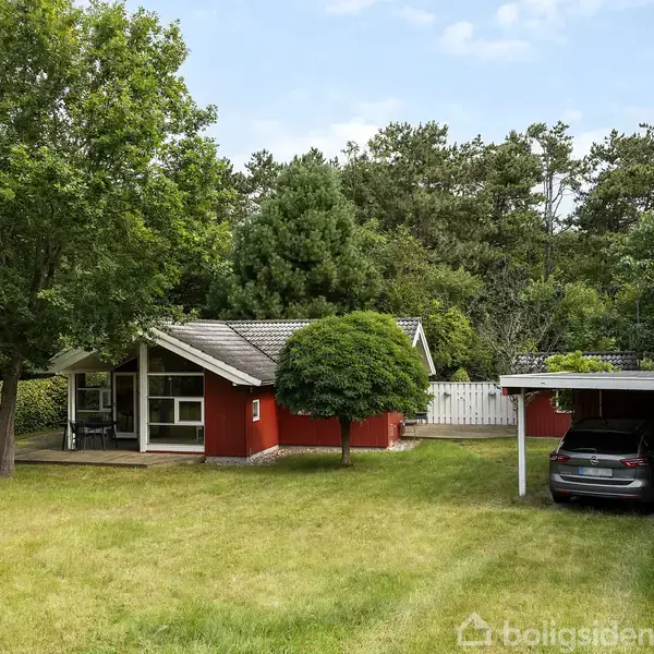 Rødt sommerhus står stille omgivet af grønne træer og græsplæne. Til højre parkeres en bil under en carport. Boligen har store vinduer og en grusbelagt indkørsel.