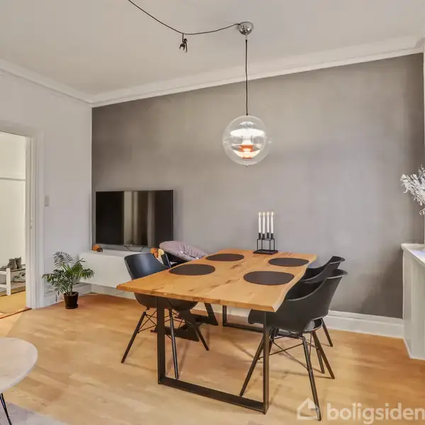 En spisebordssæt med stole står på et trægulv, oplyst af en hængelampe, i en lys stue med grå væg, vindueskarm og fjernsyn i baggrunden.