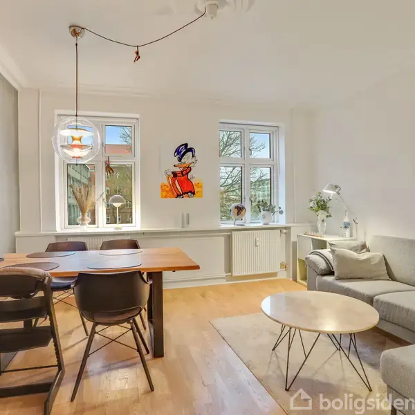 En lys stue med en grå sofa på højre side, et træspisebord og fire stole til venstre, belyst af en hængelampe. Vinduerne har udsigt til træer. En væg har en farverig illustration.