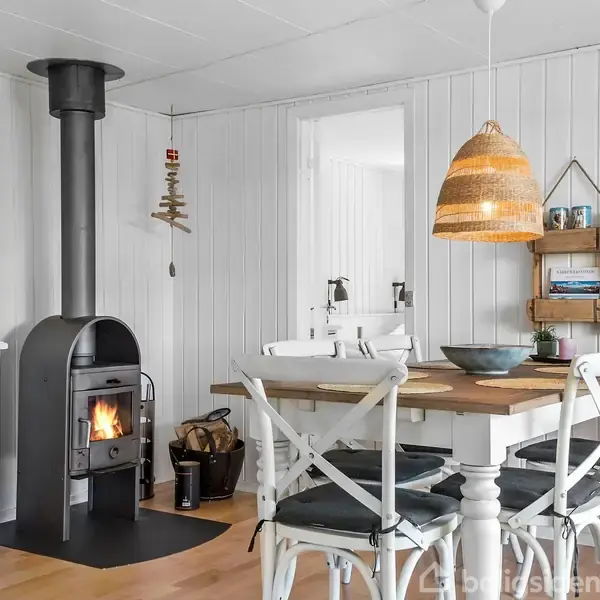 Brændeovn brænder i hjørnet af et lyst, skandinavisk køkken med hvide vægge. Et træbord med fire stole og en flettet lampe over bordet fuldender rummet.