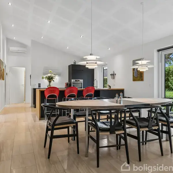 Spisebord står i moderne køkken-alrum, omgivet af sorte stole og minimalistisk indretning. Store glasdøre åbner ud til en frodig have. Tre røde stole står ved køkkenøen i baggrunden.