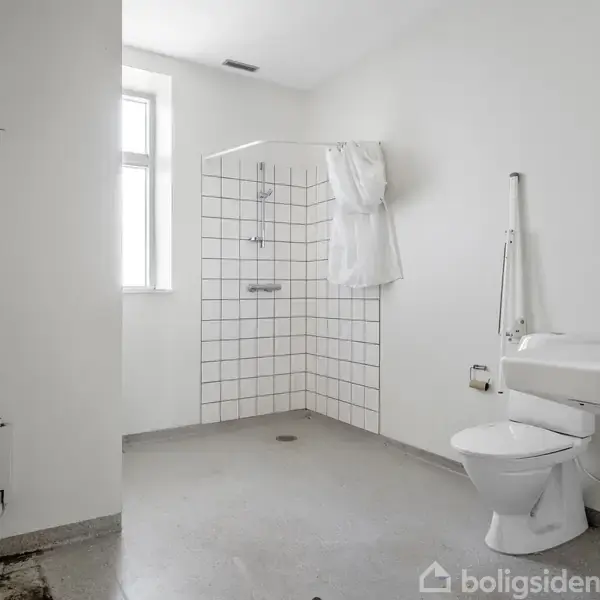 Et rummeligt badeværelse med toilet og håndvask til højre. Et bruseområde med kvadratiske fliser til venstre, placeret i en ren, lys ramme. Der er et vindue i baggrunden.