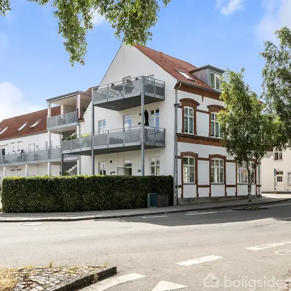 En hvid bygning med rødt tag står ved en vejkurve, omgivet af træer og grønne buske, med altaner på siden. Det er en solrig dag i et bymiljø.