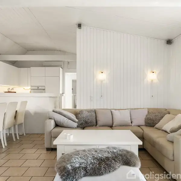 En lys stue med en stor beige hjørnesofa fyldt med puder. Et tæppe er draperet over sofabordet. Åben forbindelse til et hvidt køkken med spisebord og stole.