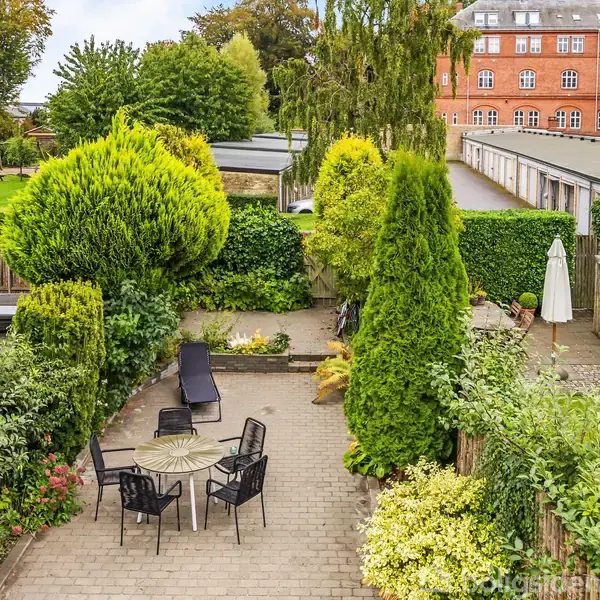 En terrasse med havemøbler står på en flisebelagt gård omgivet af grønne buske og træer. I baggrunden ses et rødt murstenshus og flere parkeringspladser.