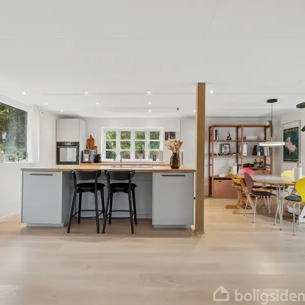 Et åbent køkken med en kogeø med barstole, omgivet af spisestue med farverige stole og hylder i baggrunden. Store vinduer giver rigeligt lys ind.