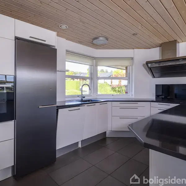 Hvidt køkken med sorte bordplader, omfatter indbyggede skabe, køleskab og ovn. Stort vindue giver udsigt til grøn have. Loftet er træbeklædt, og gulvet flisebelagt.