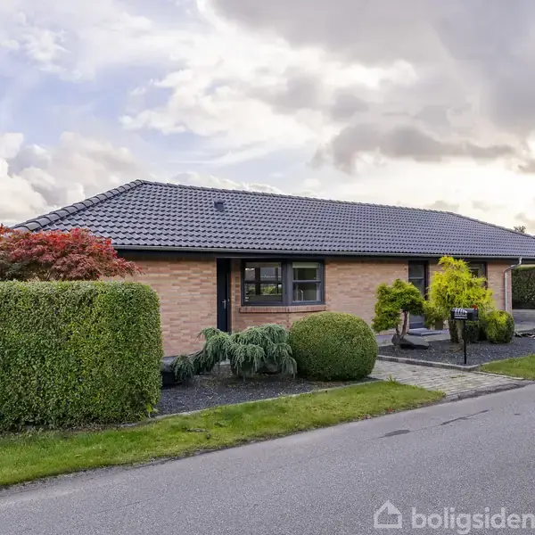 En enkeltstående etplanshus af mursten med sort tegltag står på en velplejet grund langs en stille vej. Haven er prydet med buske og træer, der giver et hyggeligt udtryk.