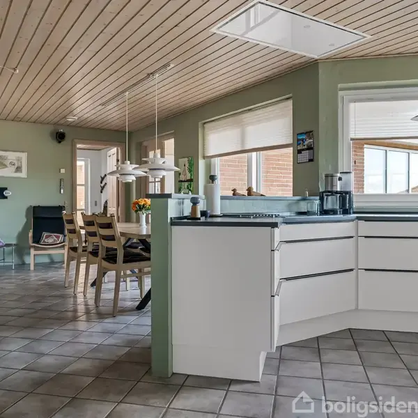 Køkken med hvidt hjørneskab, glasbord og spisebord med træstole i forgrunden. Hængelamper over bordene. Store vinduer giver naturligt lys, og der er lysegrå flisegulv og træloft.