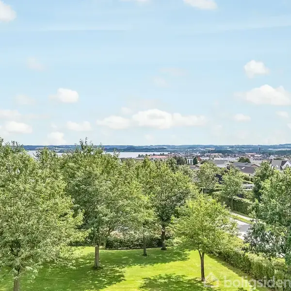 Træer står roligt i en grøn park med en udsigt over et boligområde og fjerne bakker under en klar, blå himmel.