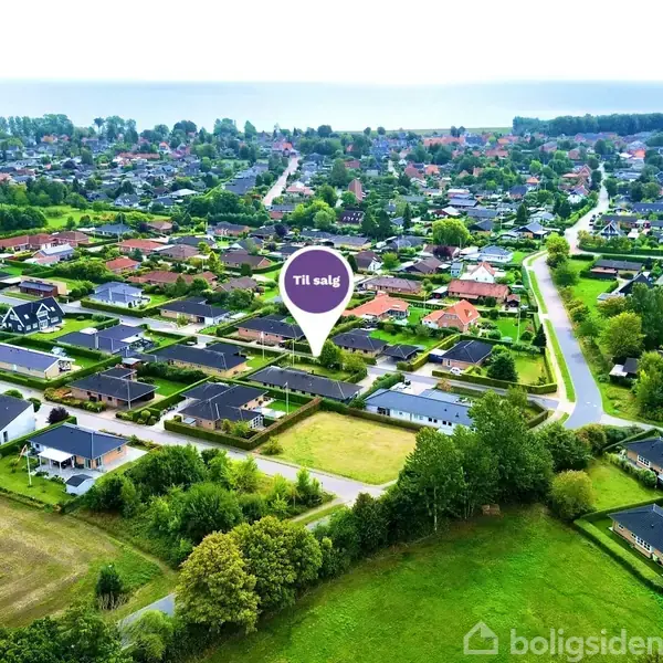 Droneperspektiv over et boligområde med huse og grønne områder. En markør peger på et hus "Til salg". I baggrunden ses en kystlinje og havet.