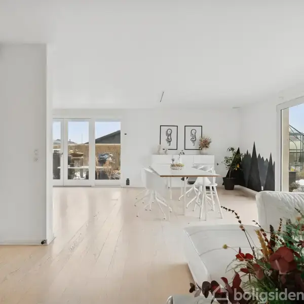 Stue med hvid sofa og blomster i forgrunden, spisebord bagved. Lysindfald fra store vinduer. Moderne indretning med kunst på væggen og planter i hjørnet.