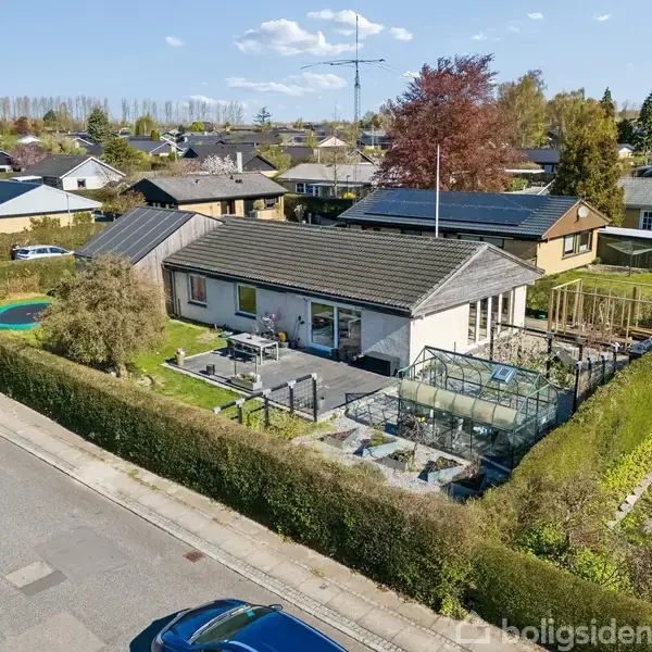 En enkeltstående etplanshus står på en velplejet grund med tilhørende drivhus og terrasse. Huset er omgivet af en hæk, på en stille villavej med flere nabohuse.