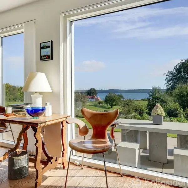 Bord med stol står i et rum med store vinduer, der giver udsigt til en grøn have og en sø i baggrunden. Lyset strømmer ind, skaber en lys, indbydende atmosfære.