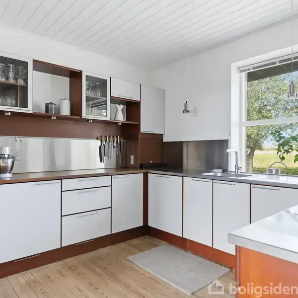 Et moderne køkken med hvide og brune skabe. En knivholder hænger på væggen, og der er en stor vinduesudsigt til grønne marker. Trægulv og hvide loftpaneler.