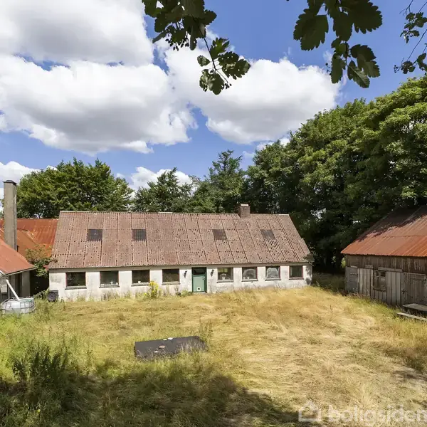Gårdskompleks med tre bygninger i en gårdsplads. Bygningerne har røde og grå tage. Tørt græs dækker jorden. Omgivet af træer og blå himmel med skyer. Ingen tekst i billedet.